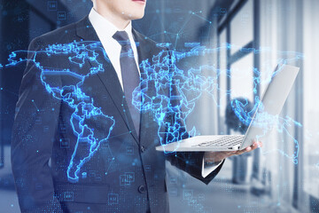 Attractive young european businessman using laptop with glowing blue map hud screen and digital data concept on blurry office interior background. Info, news and planet. Double exposure.