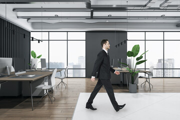 Attractive thoughtful young european businessperson walking in wooden and concrete office interior with furnituire and window with city view. CEO, executive and consulting concept.