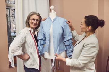 Senior fashion designer smiles posing with a dummy wearing blue shirt from new collection while her colleague tailor takes measurements and works on improvement details of new garment