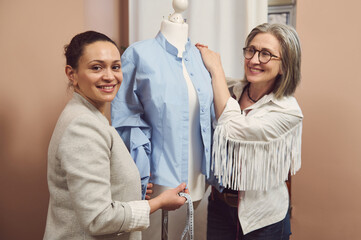 Multiethnic pretty female fashion designer with measuring tape in hand smiling while looking at camera against background of defocused tailor leaning on mannequin with shirt from new collection