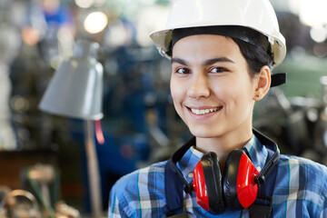 Portrait of smiling beautiful young lady worker in hardhat wearing red ear protectors on neck,...