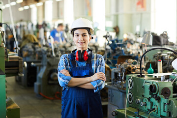 Happy attractive young lady worker in overall standing against industrial machines and smiling at...