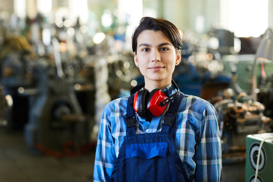 Content attractive masculine woman with short hair wearing ear protectors on neck standing at factory shop and looking at camera
