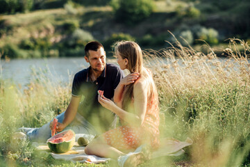 Young couple in love on summer picnic with watermelon. Loving couple sitting by the river, talking, smiling, laughing, eating watermelon