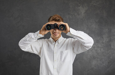 Portrait of happy young man searching for something, holding optical zoom binoculars and looking forward standing against grey studio background. Surveillance, job hunting, making discoveries concept