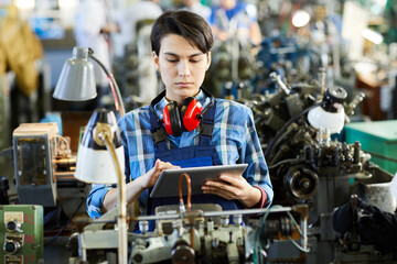 Serious concentrated busy young female factory employee with short black hair wearing ear protectors on neck using tablet while synchronizing it with modern machine