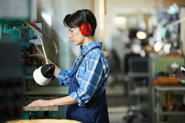 Serious skilled woman machinist in ear protectors and safety goggles pushing button while operating...