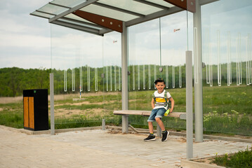 A cute six year old preschooler is sitting at the bus stop with a backpack waiting for the bus.