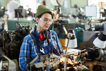 Content tough girl in beanie hat and safety goggles standing at modern lathe machine at factory and...