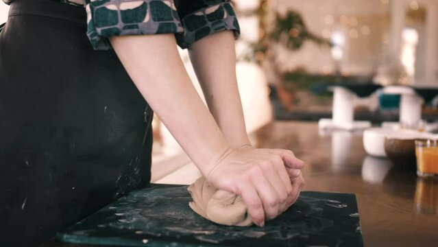 Female hands crumple and roll out piece of clay on table, front view, closeup. Potter prepares clay for processing on table.