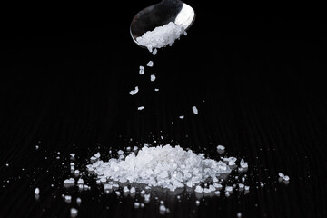 Coarse-grained sea salt is poured into a pile from a spoon against the background of a black table....