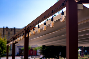 textile awning in city street cafe wooden frame gazebo with garland of strings of retro edison light bulbs glow with warm light on touristic summer day close-up cityscape, nobody. - obrazy, fototapety, plakaty
