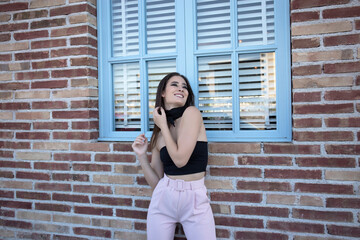 Happy young woman in fashionable outfit smiling and looking at camera while leaning on the brick wall by a window in the city street