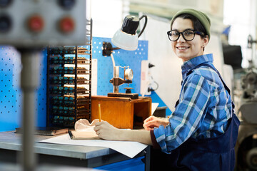 Portrait of cheerful excited attractive female watchmaker in glasses and beanie hat sitting at table and examining calculations at workplace