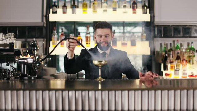 Competent barman in black suit standing behind bar counter and cleaning shiny golden wine glass with hot steam from coffee machine. Concept of restaurant service and hygiene.