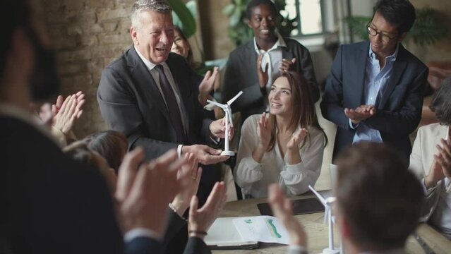 young coworkers clapping hands to business boss holding a windmill turbine in hand - celebration of the success of the project and teamwork
