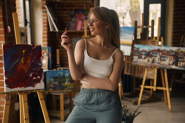 Portrait of a talented female artist working on a modern abstract oil painting, gesturing in broad strokes with a brush. Large art workshop in sunlight
