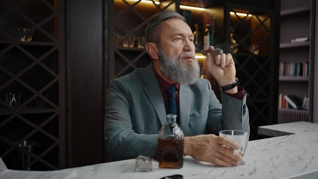 Thoughtful mature businessman enjoying cigar, relaxing with glass of whiskey at bar counter, slow motion, empty space