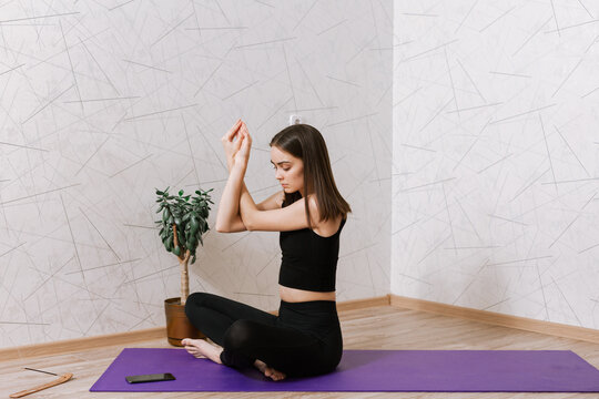 Fit young woman sitting in Eagle pose or Garudasana and doing yoga while watching online video lesson during remote session via smartphone at home 