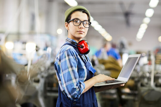 Serious confident female engineer in beanie hat and eyeglasses standing in industrial shop and using laptop while looking at camera, woman in industry concept