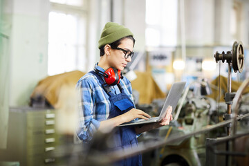 Serious busy hipster girl in workwear standing in factory shop and using laptop while examining...