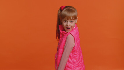 Funny joyful sincere little girl 6 years old in vest making playful silly facial expressions and grimacing, fooling around, turning, smiling. Young toddler child isolated alone on orange background