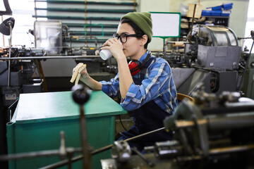 Content relaxed young woman in workwear drinking takeout coffee and eating sandwich at factory...