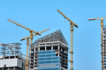 Construction of two modern skyscrapers with glasses with yellow tower cranes along the buildings with another buildings under construction around in bright sunny weather against a clear blue sky