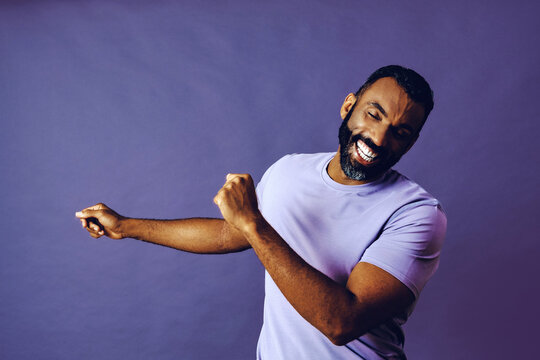 Portrait Of A Successful Man Dancing With A Beard And A Blue T-shirt Celebrating With Arm Up On A Purple Background Studio