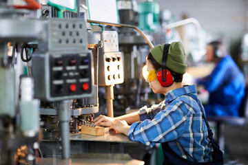 Serious busy hipster female machine operator in beanie hat adjusting metal part under press of...