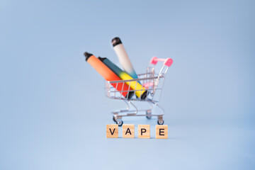 A device for vaping in a mini shopping cart on a blue background and next to the wooden letters of the word vape. the concept of vaping electronic cigarettes.