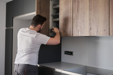 Young argentinian man installing cabinet in the kitchen using a screwdriver. Assembling modern kitchen furniture. - obrazy, fototapety, plakaty