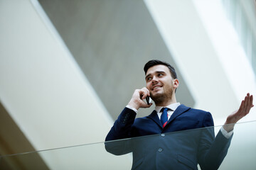 Positive confident young male entrepreneur in formal suit standing in lobby and gesturing hand in puzzlement while discussing work issues on phone