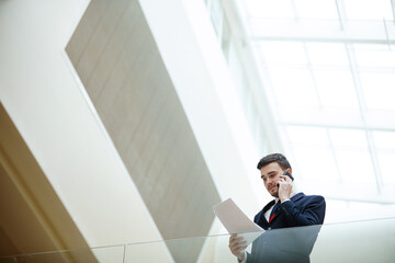 Below view of serious handsome young male manager in suit standing in modern lobby and analyzing papers while working with client on phone