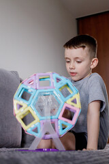 A boy in a gray T-shirt sits on a bed near a white wall and plays with a Ferris wheel, which he designed with a magnetic constructor. Creativity development. Magnetic constructor