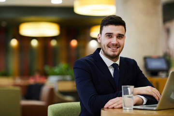 Happy successful young business manager sitting at table with laptop in restaurant and looking at camera