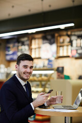 Portrait of cheerful confident young dealing manager in suit sitting at table and texting sms on smartphone while drinking water and waiting for company representative