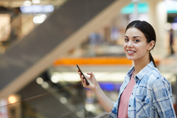 Positive beautiful girl with pretty smile wearing blue shirt standing in shopping mall and texting messages on social media while looking at camera