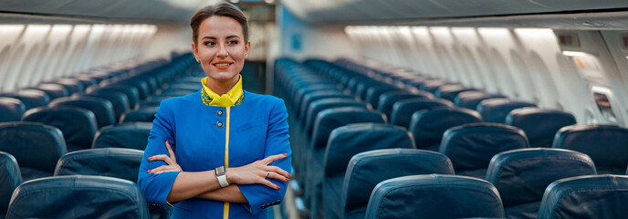 Cheerful flight attendant in air hostess uniform keeping arms crossed and smiling while standing in...