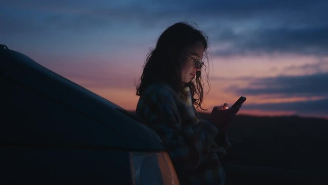 Cinematic soft view, blurry focus on young pretty millennial woman using smartphone, smiling, scrolling social media outdoors leaning on camper car on weekend, trip, vacation on colorful Porto sunset