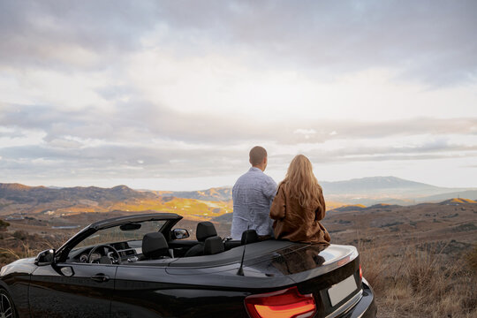 Rear of young couple in love man and woman enjoying mountains view standing near convertible car
