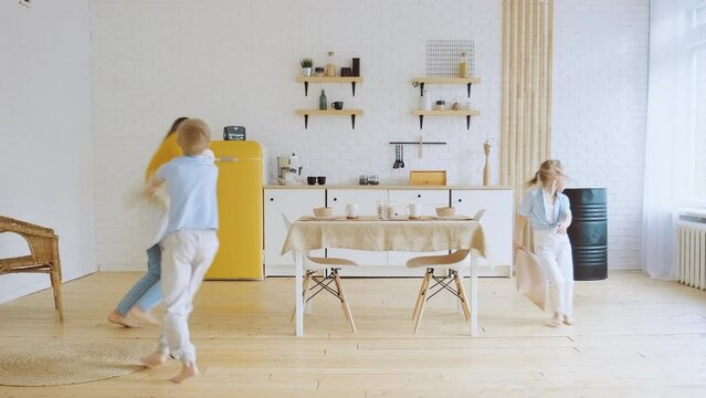 Mother and children have fun running at home around kitchen table fighting on pillow. Leisure of mom, son, daughter, true emotions. Happy merry family, parents play with small kids.