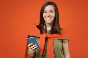 Young smiling happy woman 20s wearing khaki t-shirt tied sweater on shoulders hold in hand use mobile cell phone surfing internet isolated on plain orange background studio. People lifestyle concept.