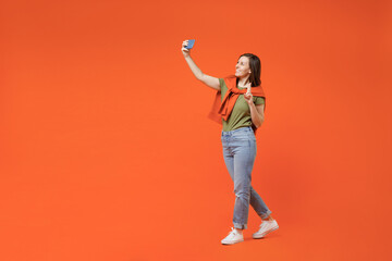 Fototapeta na wymiar Full body young smiling happy woman 20s in khaki t-shirt tied sweater on shoulders doing selfie shot pov on mobile cell phone show v-sign isolated on plain orange background. People lifestyle concept.