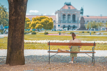 Young woman sitting on a bench and reading in the shadow under the trees in Kralj Tomislav park in zagreb, looking towards the train station.