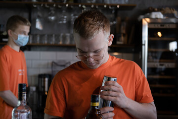 stylish young man hipster in orange t-shirt making mixing a cocktail in a dark loft cafe. alcohol drink in modern bar.