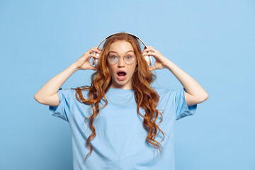 Shocked young redhead girl, student in headphones isolated on blue studio background. Human emotions, facial expression concept. Trendy colors