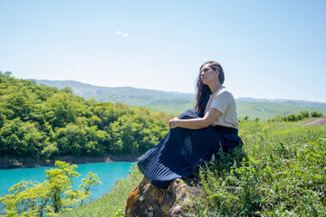 Naklejka na ściany i meble A girl in a dress sits on the shore of a blue lake in summer