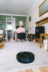 Selective focus on vacuum cleaner robot controlled by senior woman through phone settings while...