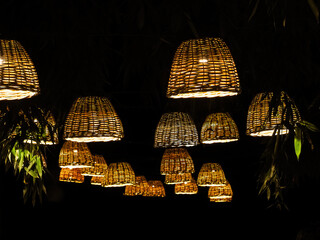 Many lamps with wicker lampshades hang in a dark bamboo park. - 508429504